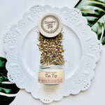 Blooming with Joy Tea Tox herbal loose leaf tea, showing dry herbs spilling from a small jar onto a white decorative plate with green leaves in the background.